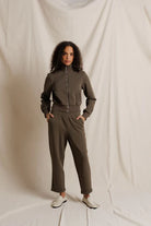 A woman modeling a matching two-piece tracksuit in a muted olive-brown color, consisting of a zip-up long-sleeve top and matching pants, standing against a draped off-white fabric backdrop.