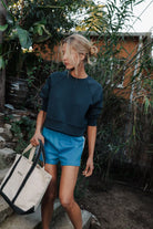 Woman in navy sweatshirt and blue shorts holding a beige tote bag in a natural setting.
