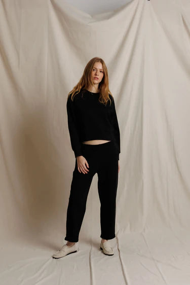 A model stands against a draped beige fabric backdrop wearing a black long-sleeve cropped top and black pants with white slip-on shoes.