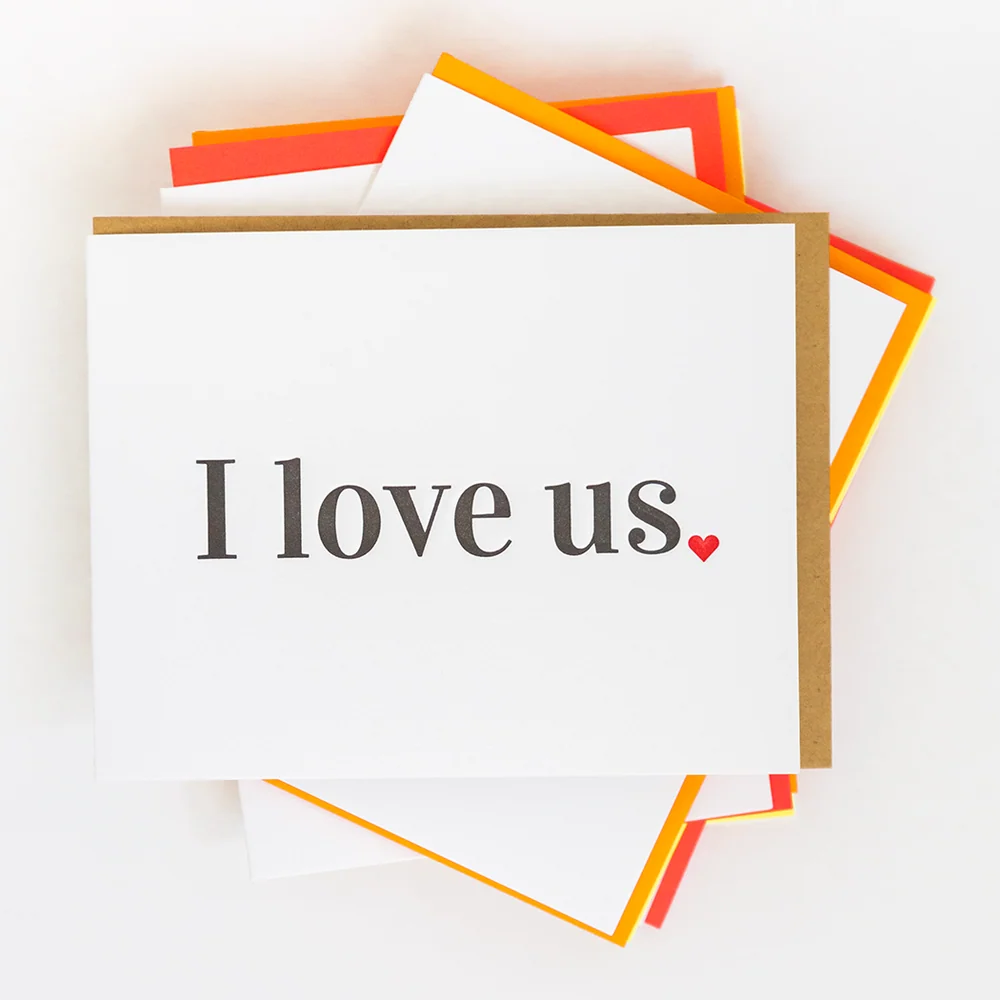 A white greeting card with the words 'I love us.' in dark gray serif font and a small red heart, stacked on top of other cards with orange and red borders on a white background.