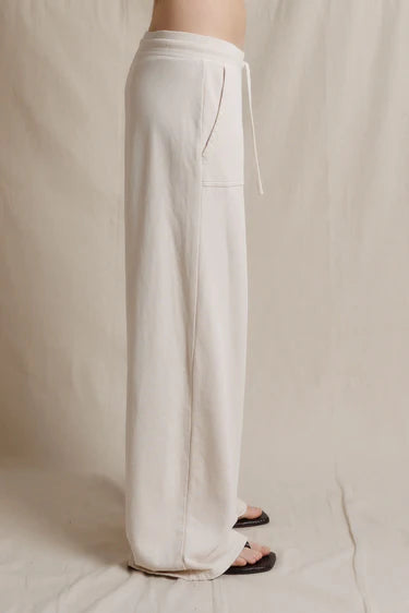 A person wearing loose, wide-leg white drawstring pants with a side pocket, shown from the waist down against a neutral beige fabric backdrop.