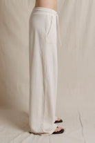 A person wearing loose, wide-leg white drawstring pants with a side pocket, shown from the waist down against a neutral beige fabric backdrop.