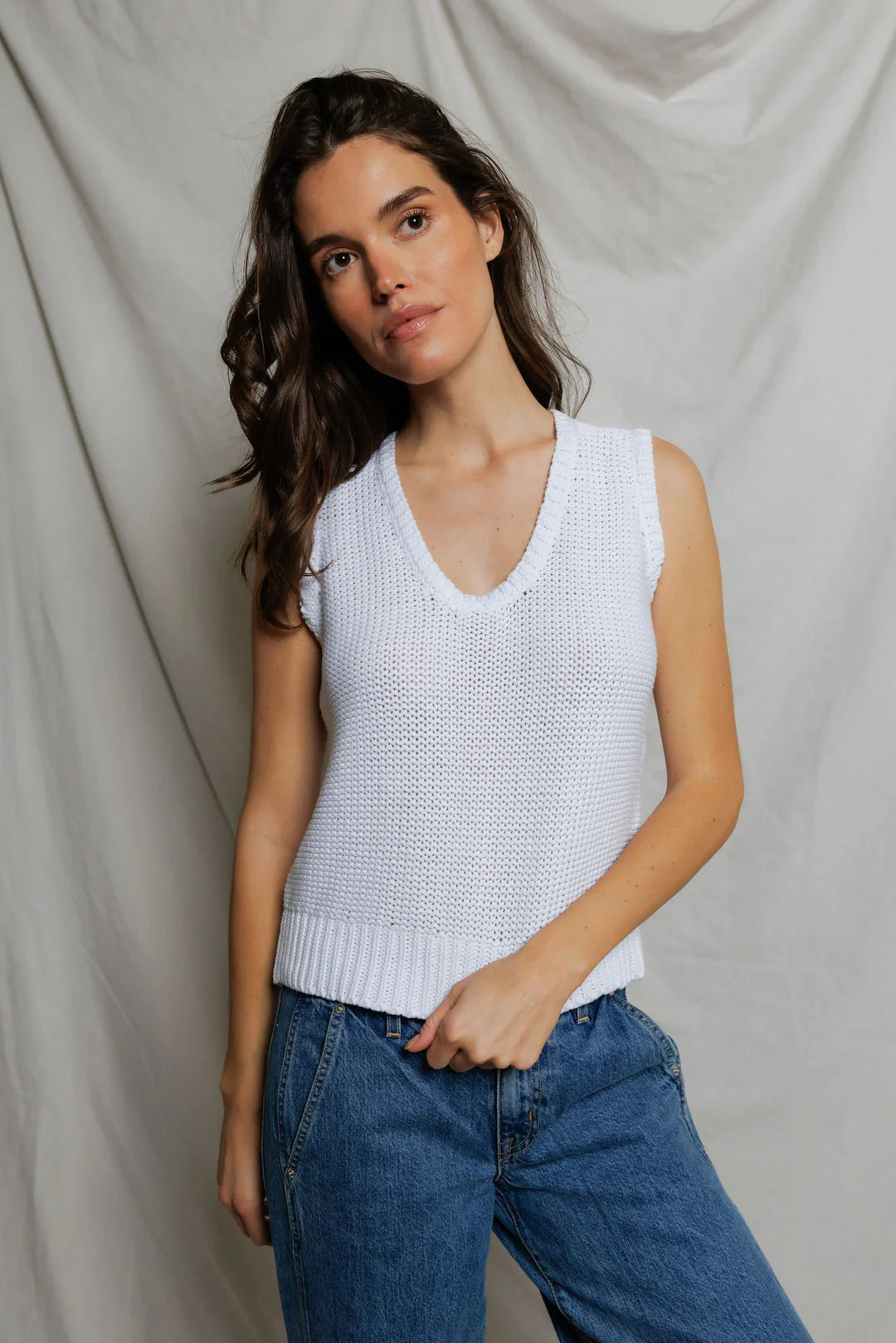 Woman wearing a white sleeveless knitted top and blue jeans against a plain background