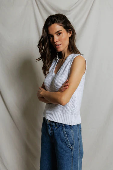 Woman wearing a light blue sleeveless knitted top and blue jeans against a plain background