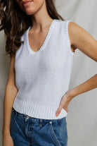 Woman wearing a white knitted sleeveless top and blue jeans against a neutral background