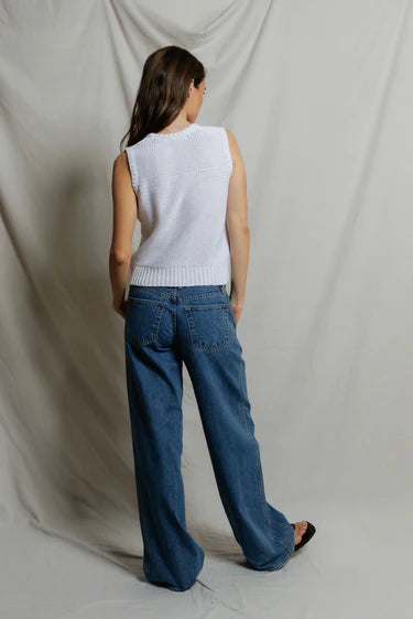 Person wearing a white sleeveless sweater and blue jeans against a plain background