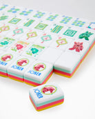 A close-up of a set of Mahjong-style tiles on a white background, with one tile in front showing the Joker symbol and pastel pink, yellow, and mint green layered edges.