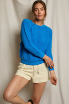Woman wearing a blue sweater and beige shorts against a neutral background
