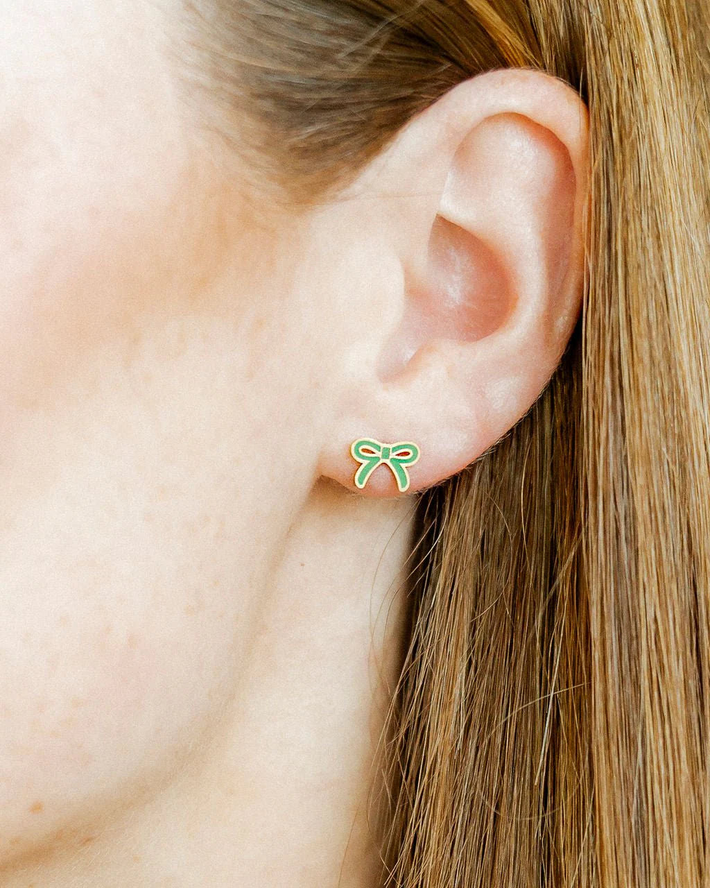 Close-up of an ear wearing a green bow-shaped earring with blonde hair.