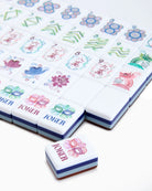 A close-up of Mahjong tiles on a white background, showing the back of several tiles with blue and brown layers and one loose tile in front labeled JOKER with pink and green floral designs.