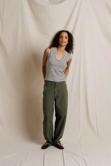 A woman stands against a draped off-white fabric backdrop wearing a sleeveless black and white striped tank top, olive green wide-leg pants, and dark flat shoes.