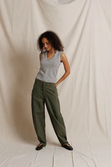 A woman modeling a sleeveless black and white striped tank top with olive green wide-leg pants, standing against a draped off-white fabric backdrop.