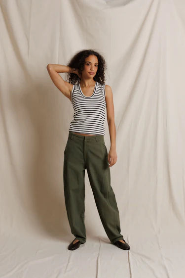A model stands against a draped off-white fabric backdrop, wearing a black and white horizontally striped tank top, olive green wide-leg trousers, and dark brown flat shoes.