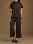 A model wearing a dark brown two-piece outfit consisting of a short-sleeve mock-neck top and matching wide-leg cropped pants, paired with dark brown loafers, against a solid warm brown studio background.