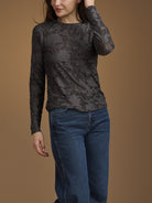 A woman wearing a dark charcoal gray long-sleeve top with an intricate floral lace pattern, paired with blue jeans, set against a solid warm brown studio background.