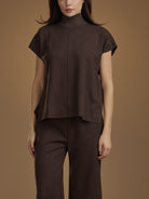 A model wearing a dark brown short-sleeve mock-neck top and matching dark brown trousers, standing against a solid warm brown studio background.