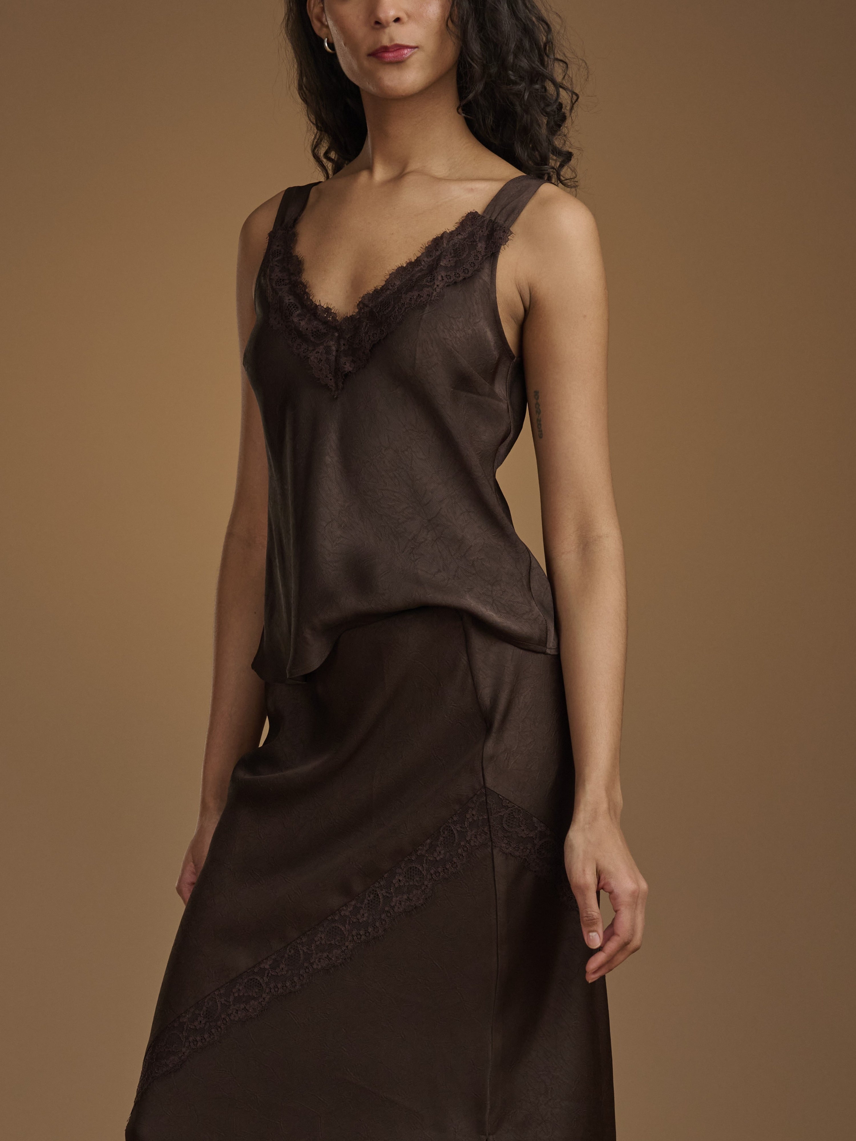 A woman wearing a dark brown satin slip dress with lace trim, photographed against a solid warm brown studio background.