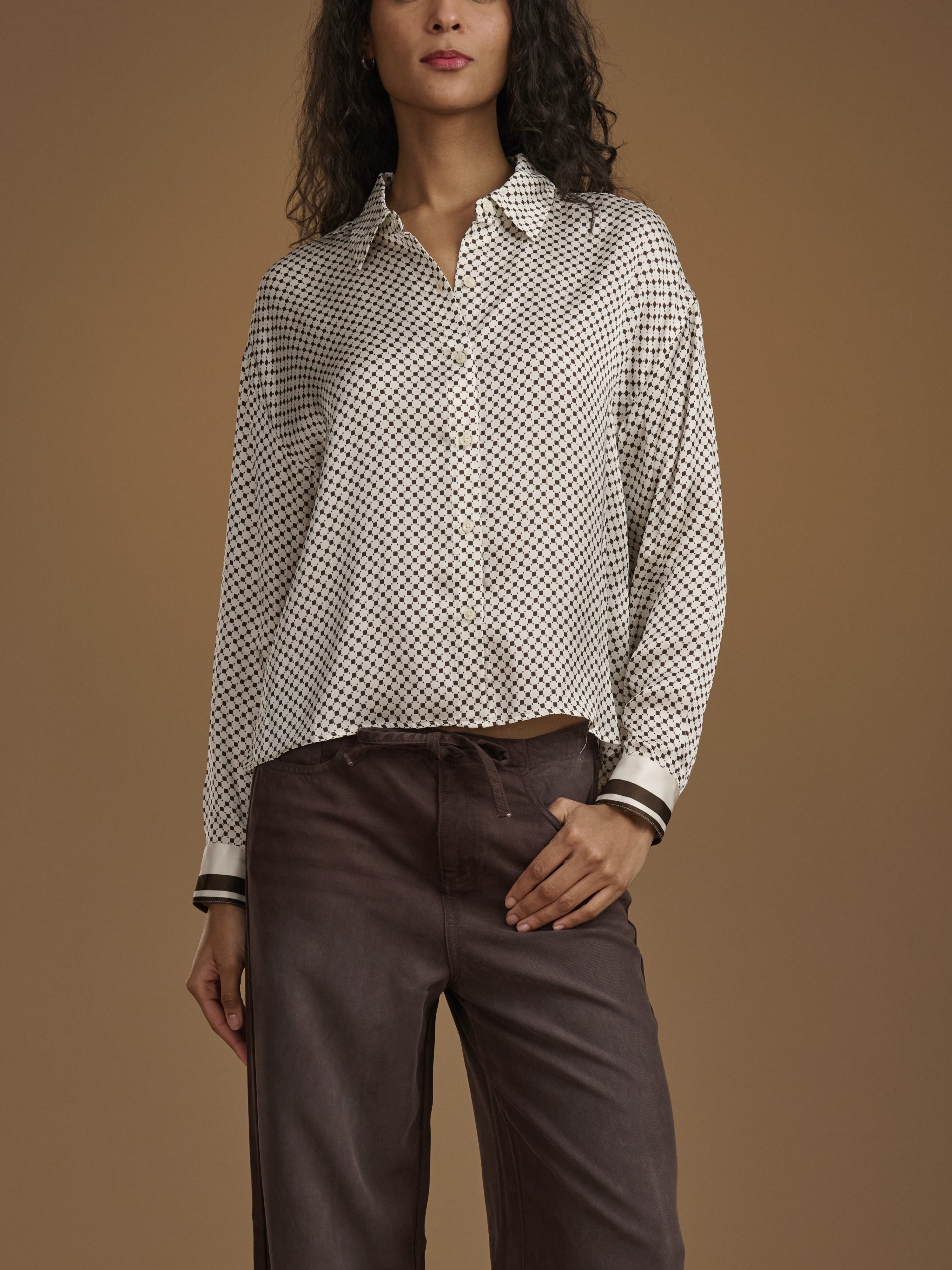 A woman wearing a long-sleeve button-up shirt with a small black-on-white geometric pattern, paired with dark brown trousers, set against a solid warm brown studio background.