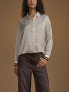 A woman wearing a long-sleeve button-up shirt with a small black-on-white geometric pattern, paired with dark brown trousers, set against a solid warm brown studio background.