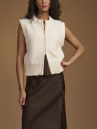 A woman models a sleeveless cream cable-knit zip-front vest over a dark brown skirt, set against a solid warm brown studio background.