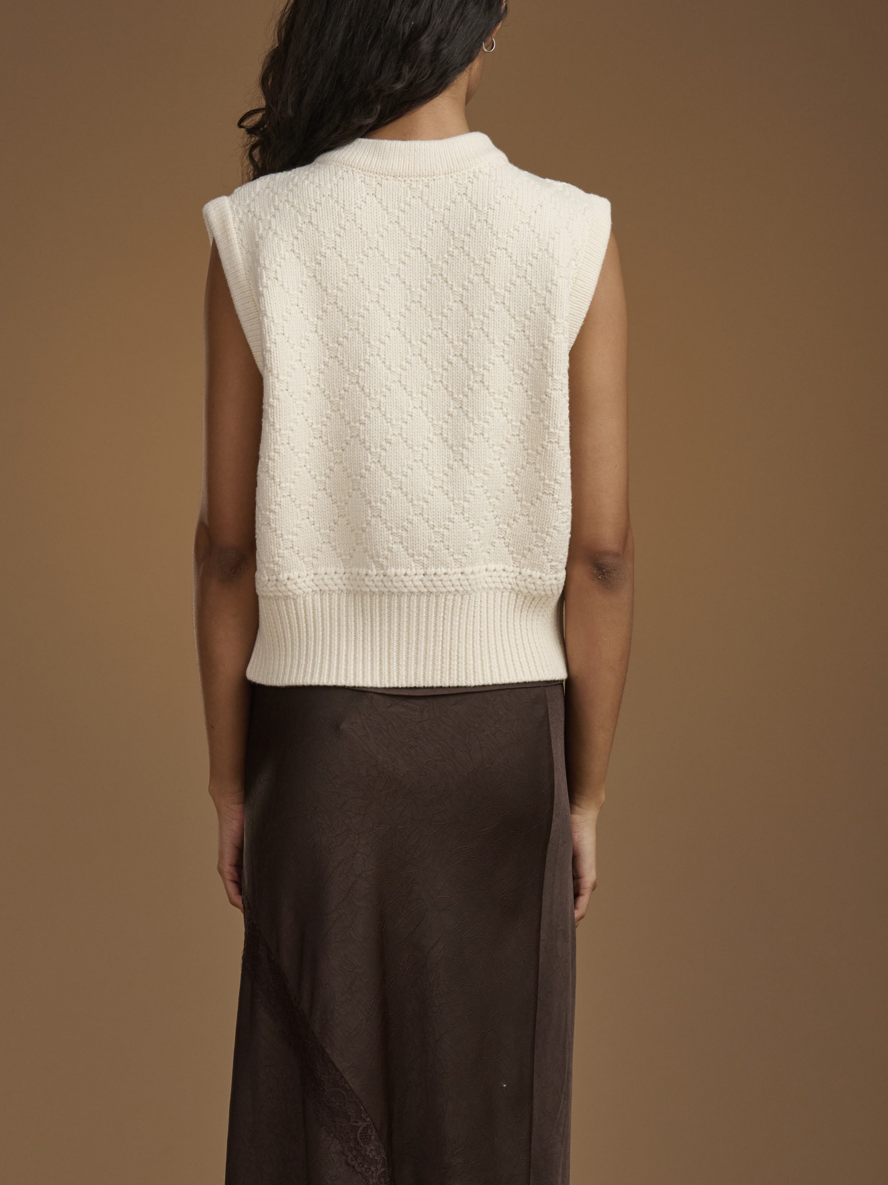 Back view of a model wearing a cream-colored sleeveless knit sweater vest over a dark brown skirt, set against a solid warm brown studio background.