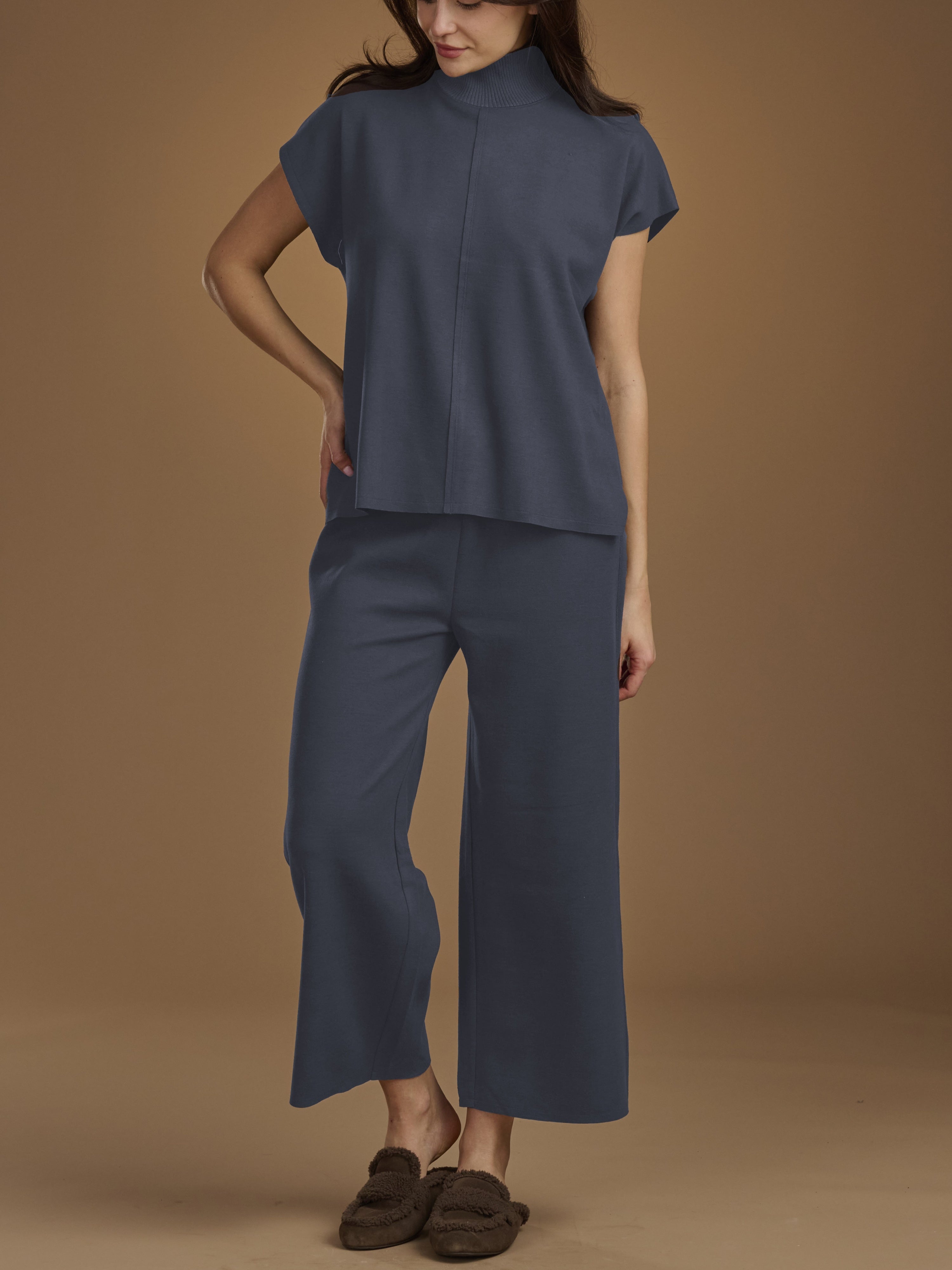 A woman modeling a matching navy blue two-piece set consisting of a short-sleeve mock-neck top and wide-leg cropped pants, paired with brown slip-on shoes against a solid tan studio background.