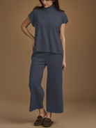 A woman modeling a matching navy blue two-piece set consisting of a short-sleeve mock-neck top and wide-leg cropped pants, paired with brown slip-on shoes against a solid tan studio background.