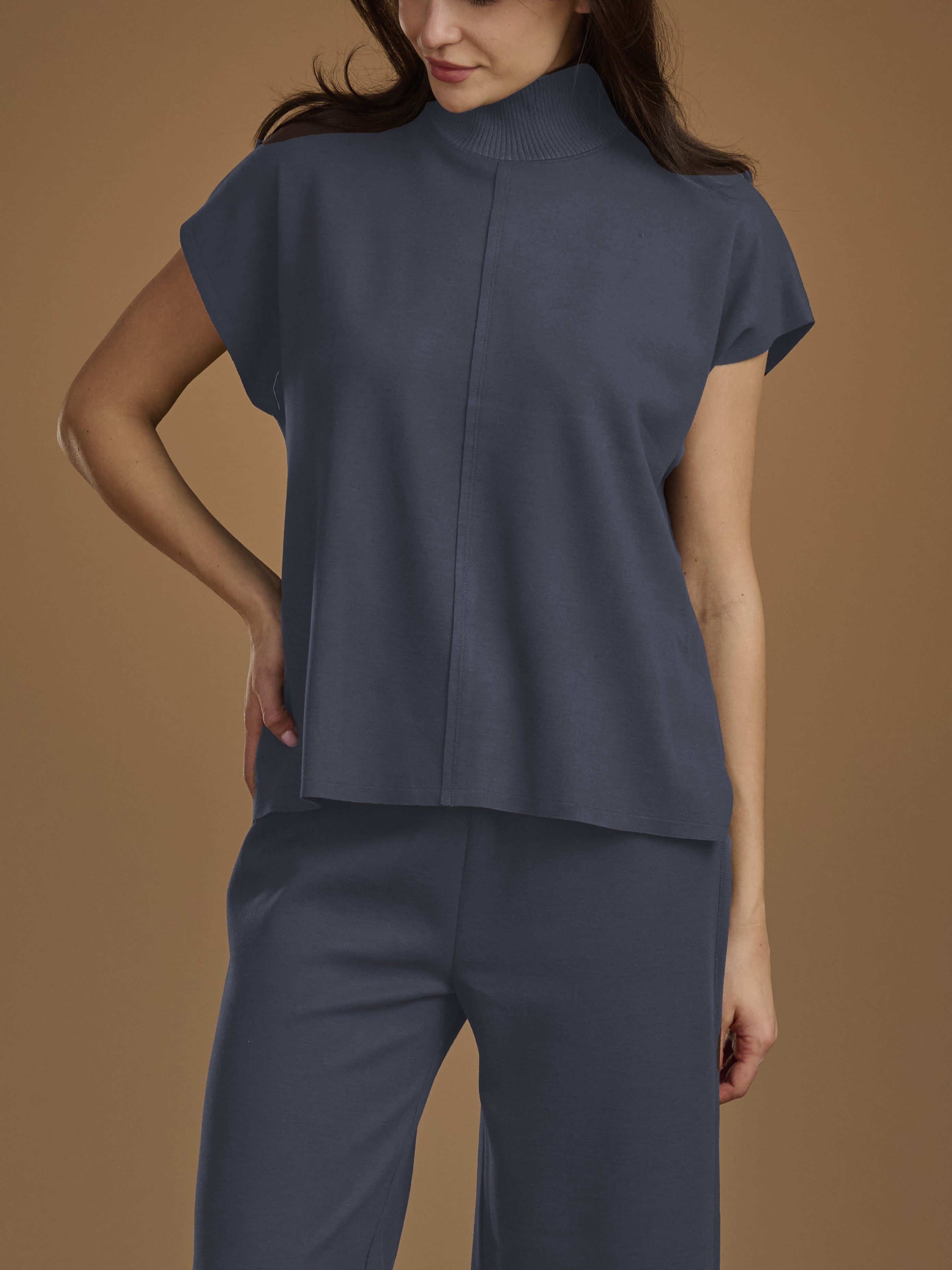 A woman wearing a slate blue mock-neck short-sleeve top and matching wide-leg trousers, posed against a solid warm brown studio background.
