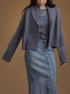 A woman wearing a slate blue open-front knit cardigan over a matching lace top and a blue denim skirt, photographed against a solid warm brown studio background.