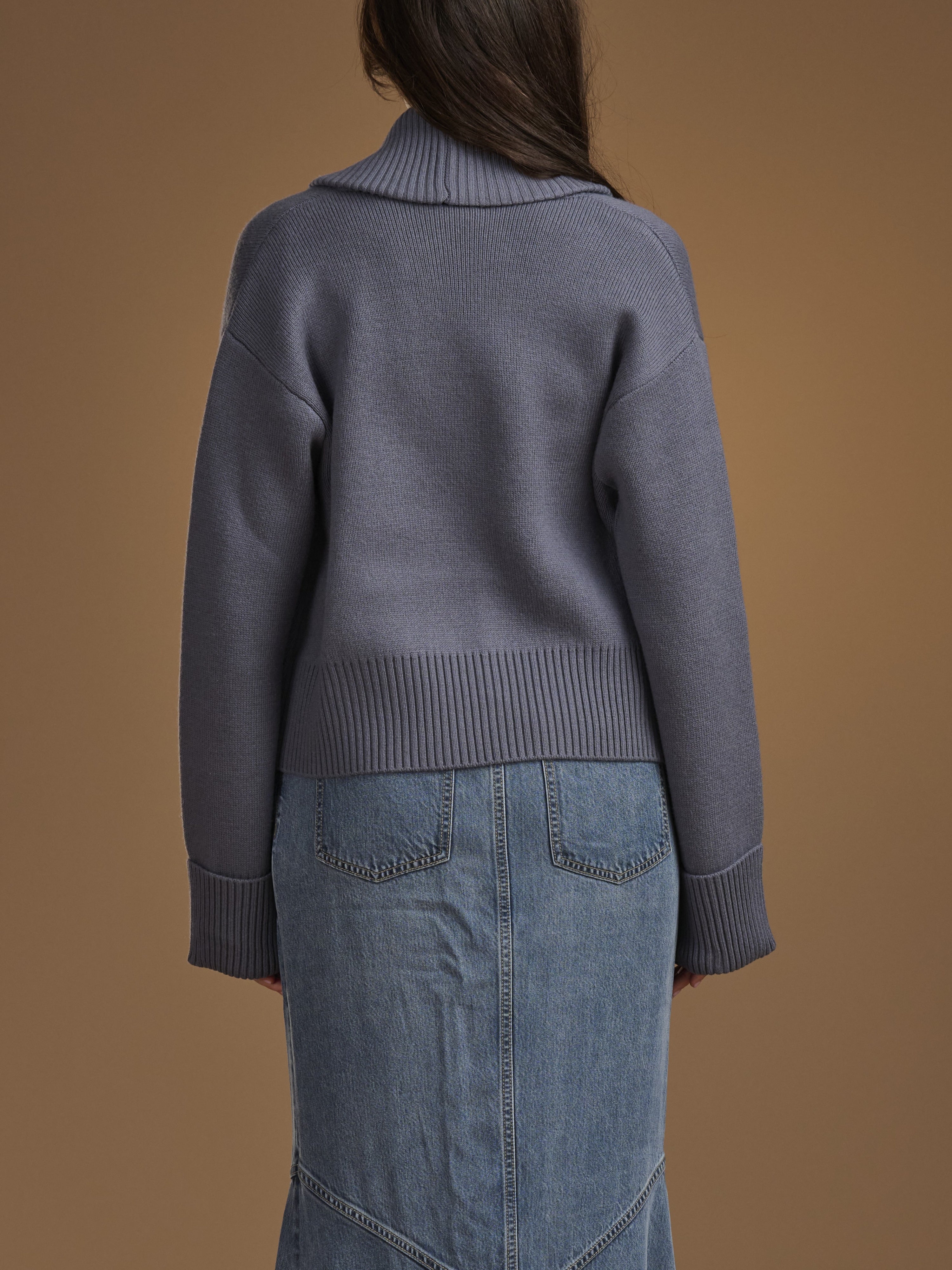 A person shown from the back wearing a slate blue-gray ribbed knit cardigan and a light blue denim skirt, set against a solid warm brown studio background.