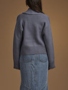 A person shown from the back wearing a slate blue-gray ribbed knit cardigan and a light blue denim skirt, set against a solid warm brown studio background.