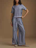 A woman models a matching two-piece loungewear set consisting of a short-sleeve cropped top and wide-leg pants, both in a blue and white geometric pattern. She is standing against a solid warm brown studio backdrop.
