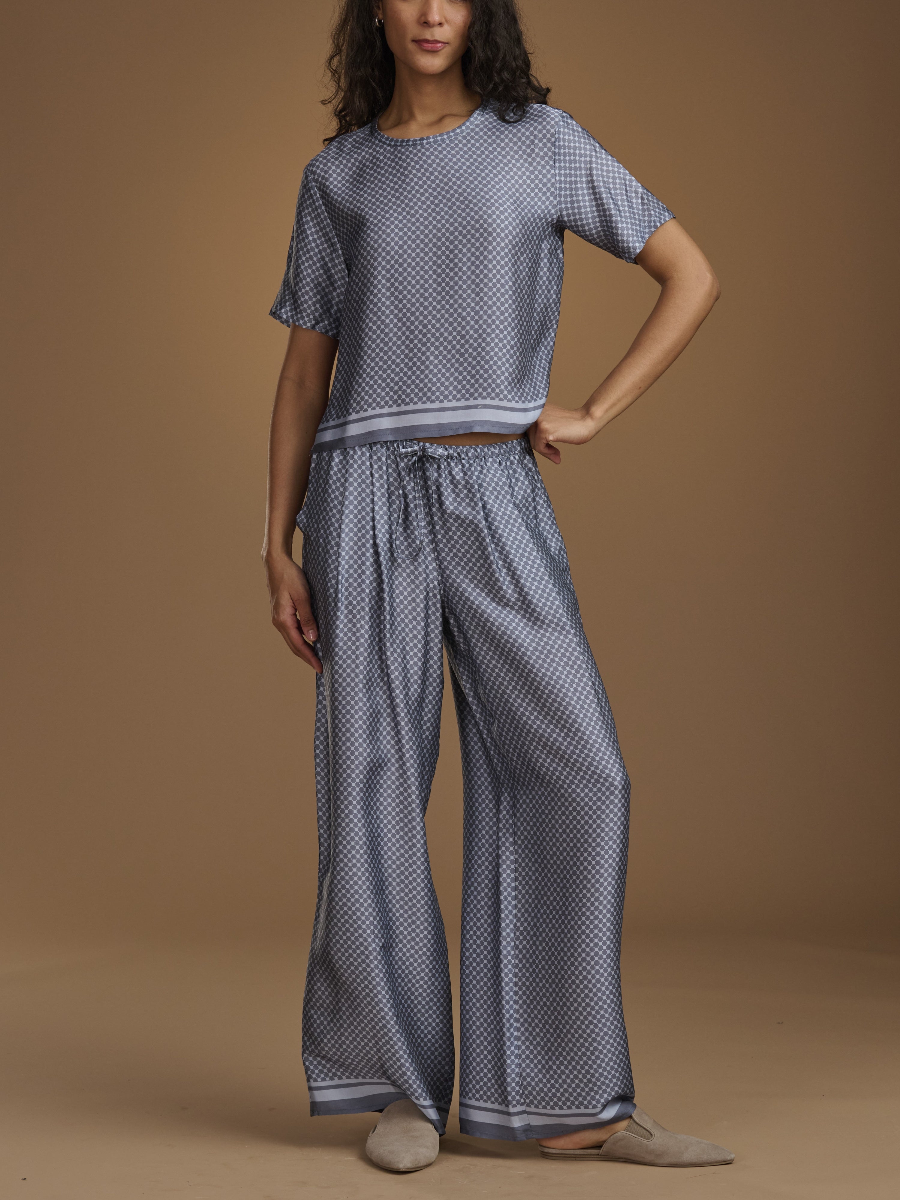 A woman models a matching two-piece loungewear set consisting of a short-sleeve cropped top and wide-leg pants, both in a blue and white geometric pattern. She is standing against a solid warm brown studio backdrop.