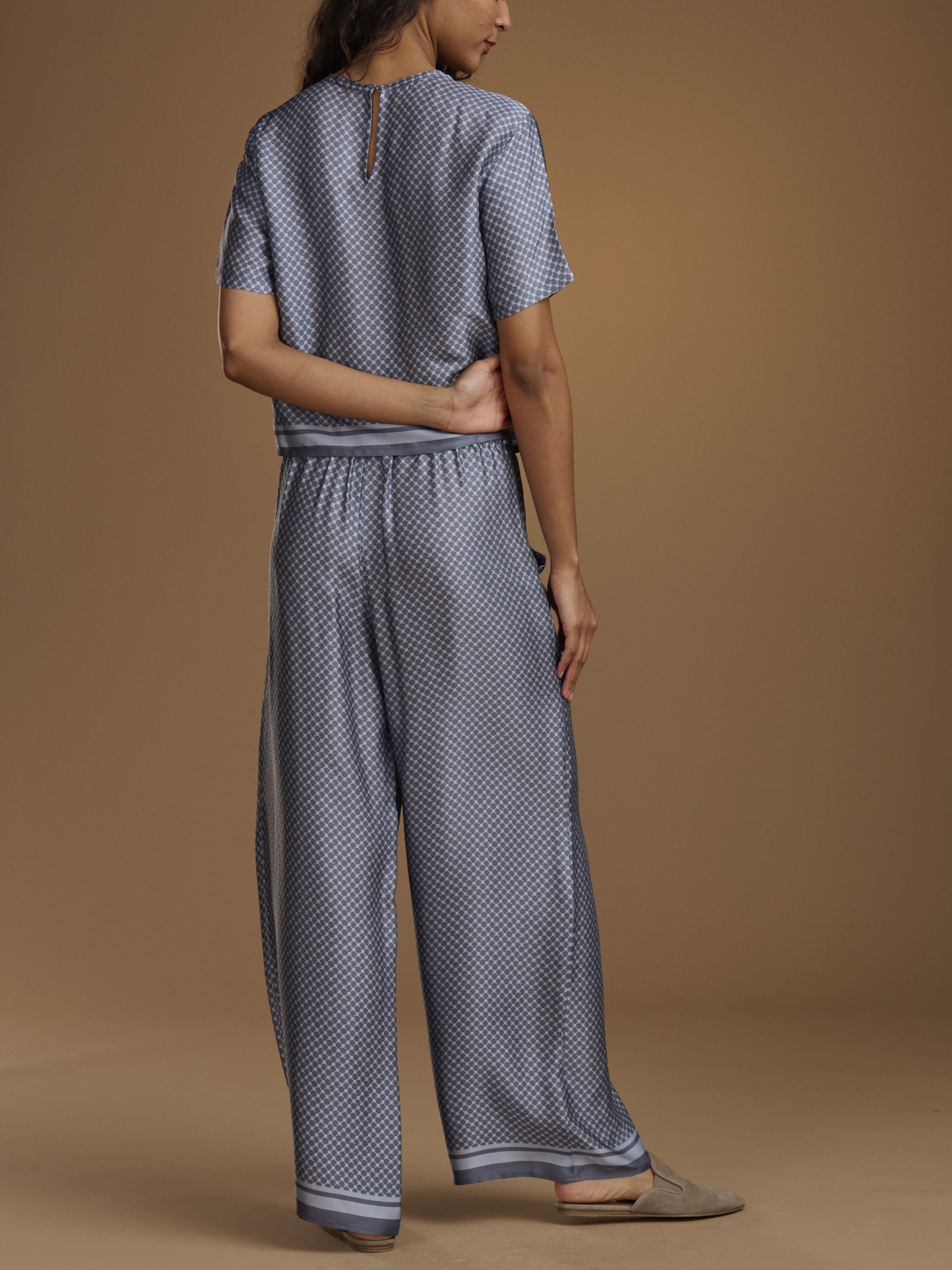 A woman shown from the back wearing a matching short-sleeve top and wide-leg pants set in a blue and white micro-pattern, against a solid tan studio background.