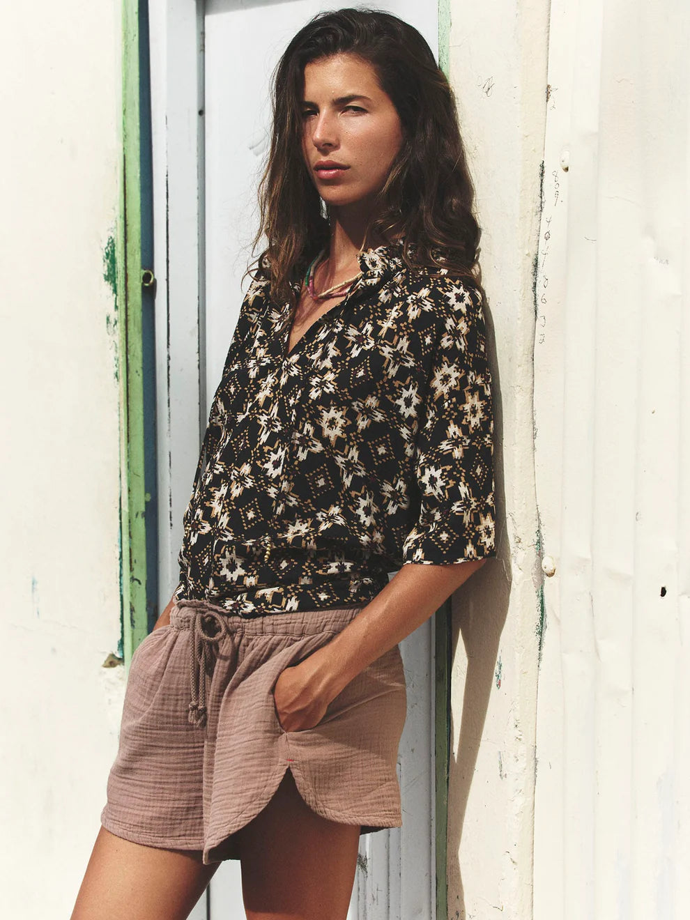 Woman wearing a floral shirt and shorts leaning against a white wall.