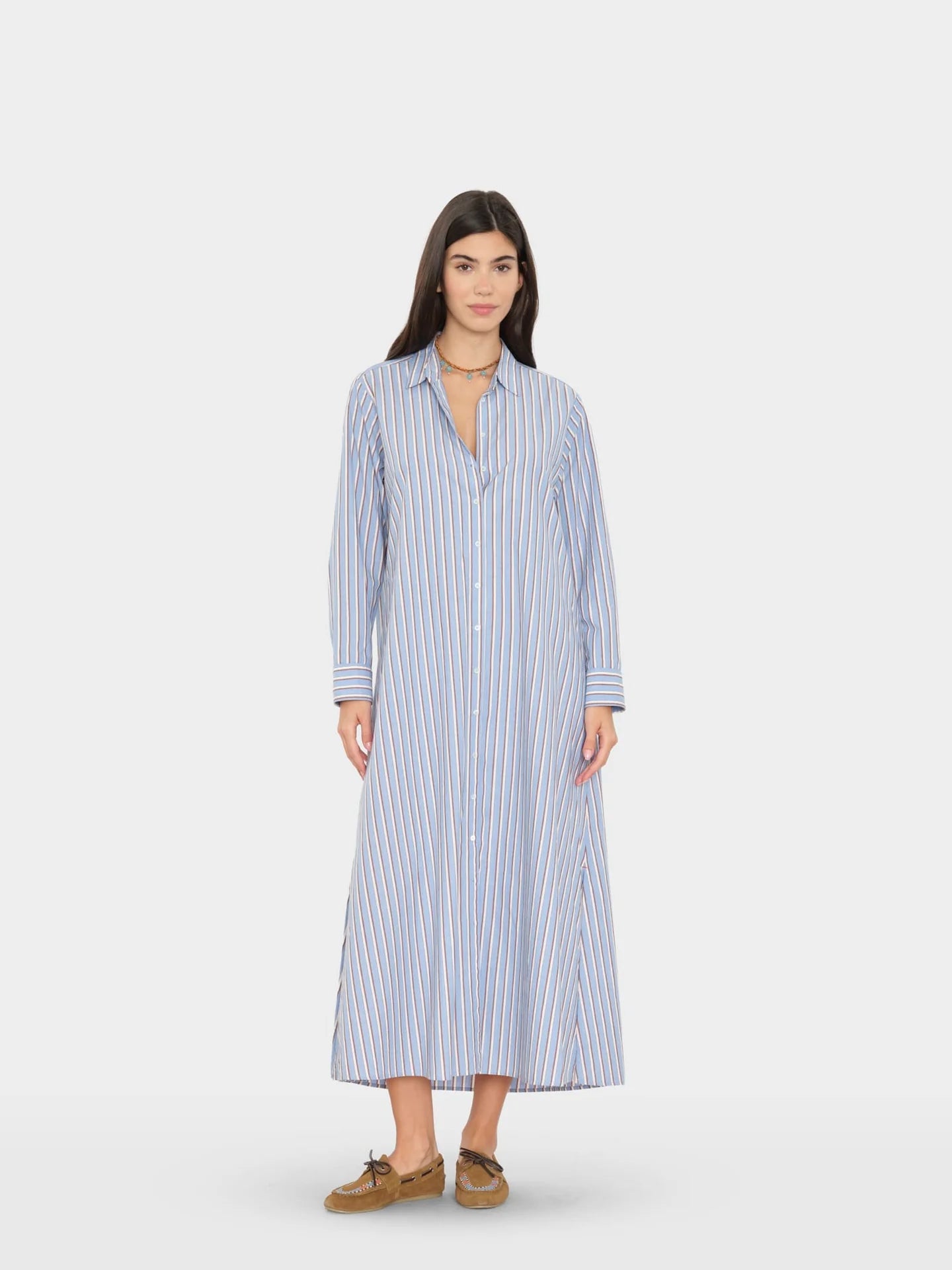 A woman modeling a long-sleeve, button-front midi dress with vertical blue and white stripes, paired with brown loafers, against a light gray studio background.