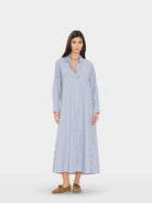 A woman modeling a long-sleeve, button-front midi dress with vertical blue and white stripes, paired with brown loafers, against a light gray studio background.