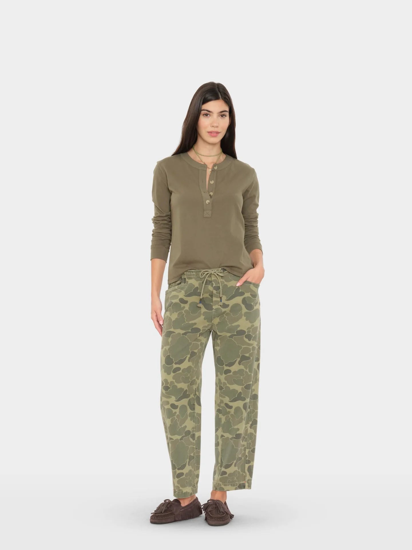 A woman modeling a matching two-piece loungewear set consisting of an olive green henley long-sleeve top and green camouflage drawstring pants, paired with dark brown loafers, against a light gray studio background.
