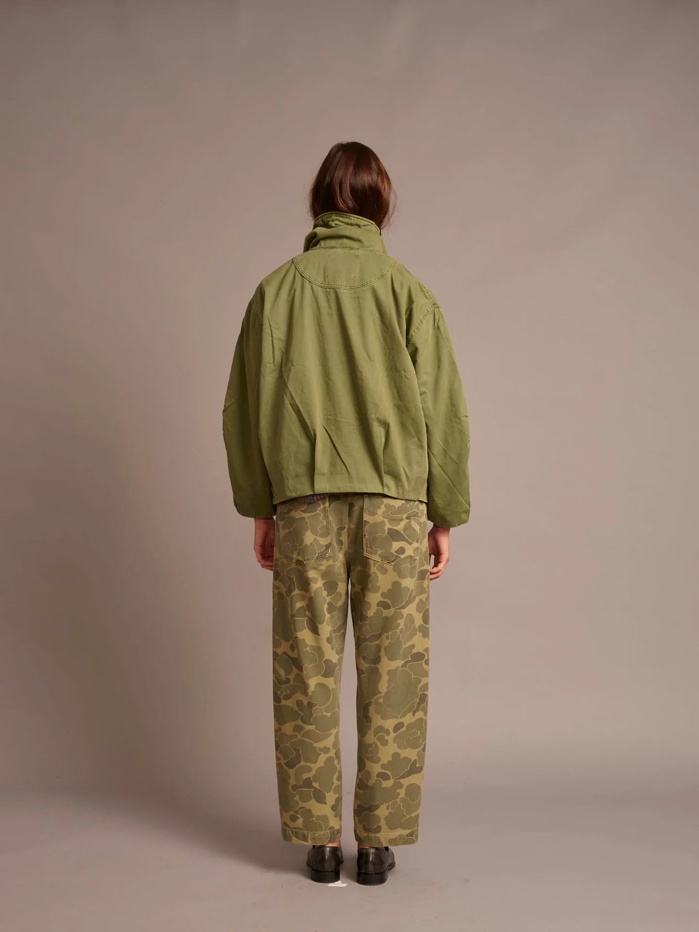 A person shown from the back wearing an olive green jacket and olive-green camouflage pants, standing against a plain warm gray studio background.