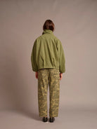 A person shown from the back wearing an olive green jacket and olive-green camouflage pants, standing against a plain warm gray studio background.