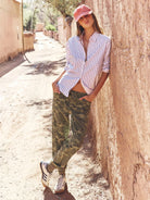 A woman leans against a sunlit stucco wall in a narrow outdoor alley, wearing a pink and white striped button-up shirt, green camouflage jogger pants, white sneakers, and a pink cap.
