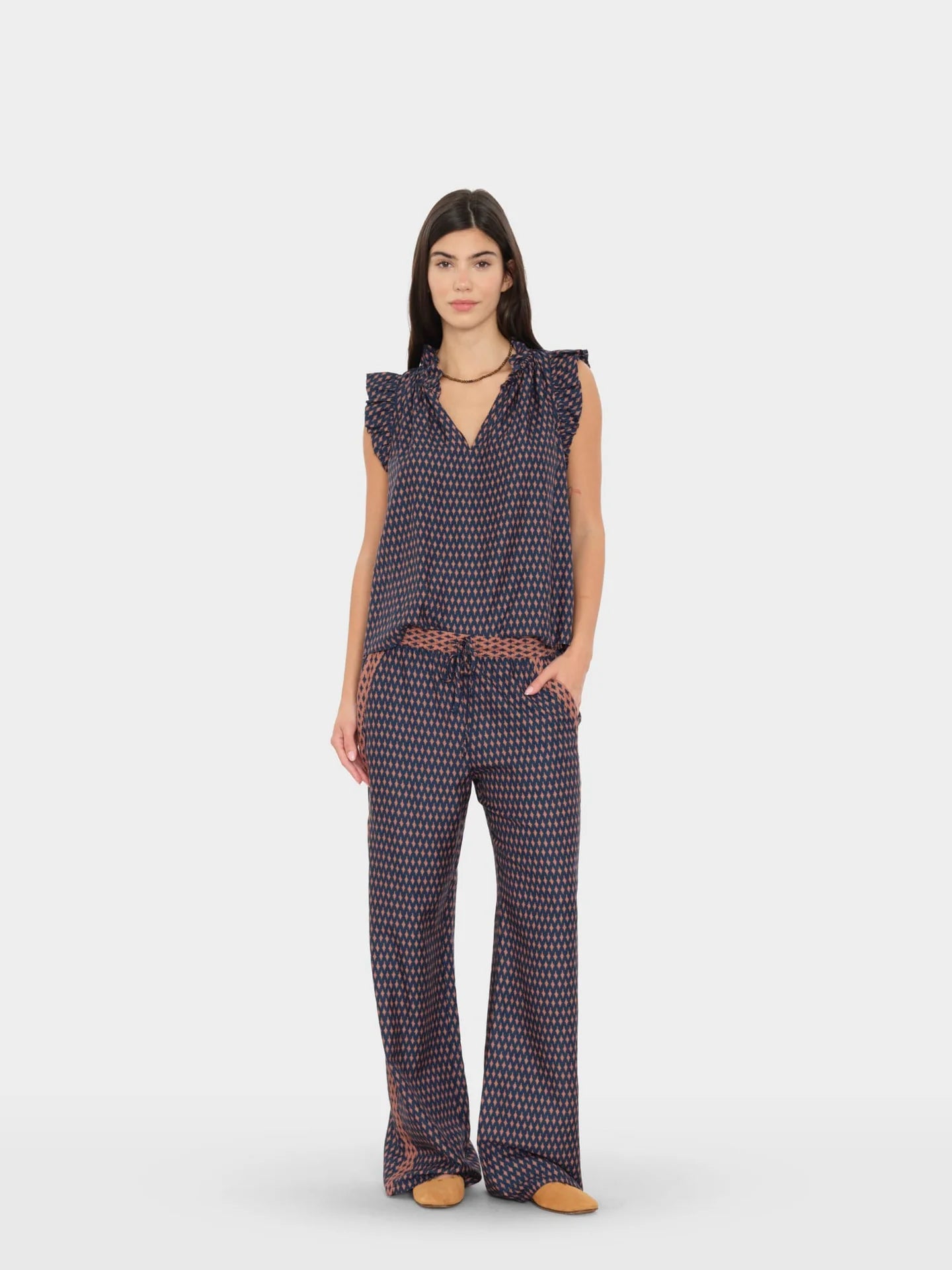 A woman modeling a sleeveless navy jumpsuit with a dense small white and orange geometric pattern, featuring ruffled shoulders, a V-neckline, side pockets, and a matching patterned belt, set against a plain light gray studio background.
