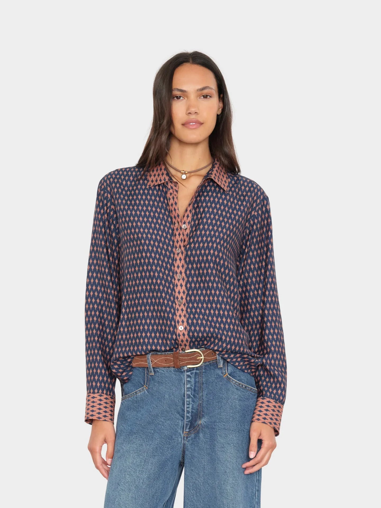 A woman modeling a long-sleeve button-up shirt with a small geometric pattern in navy and dusty pink, paired with blue jeans and a brown belt, against a light gray background.
