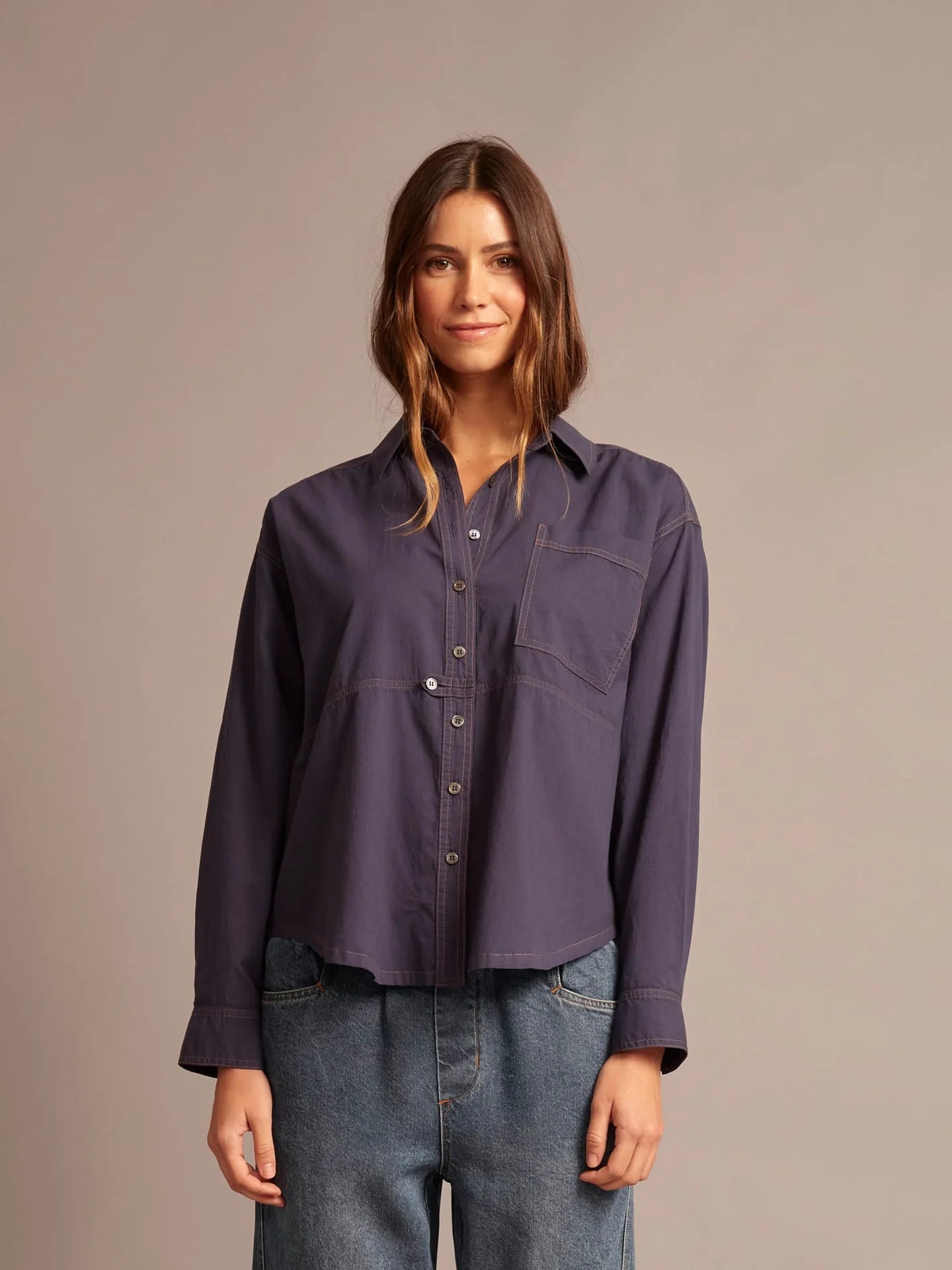 A woman modeling a dark purple, long-sleeve button-up shirt with a collar and chest pocket, paired with blue jeans, against a solid taupe background.