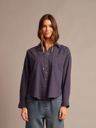 A woman modeling a dark purple, long-sleeve button-up shirt with a collar and chest pocket, paired with blue jeans, against a solid taupe background.