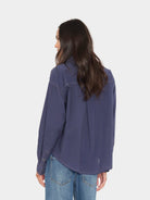A woman with long dark hair is shown from the back wearing a loose, long-sleeve blue shirt and light wash jeans against a light gray studio background.