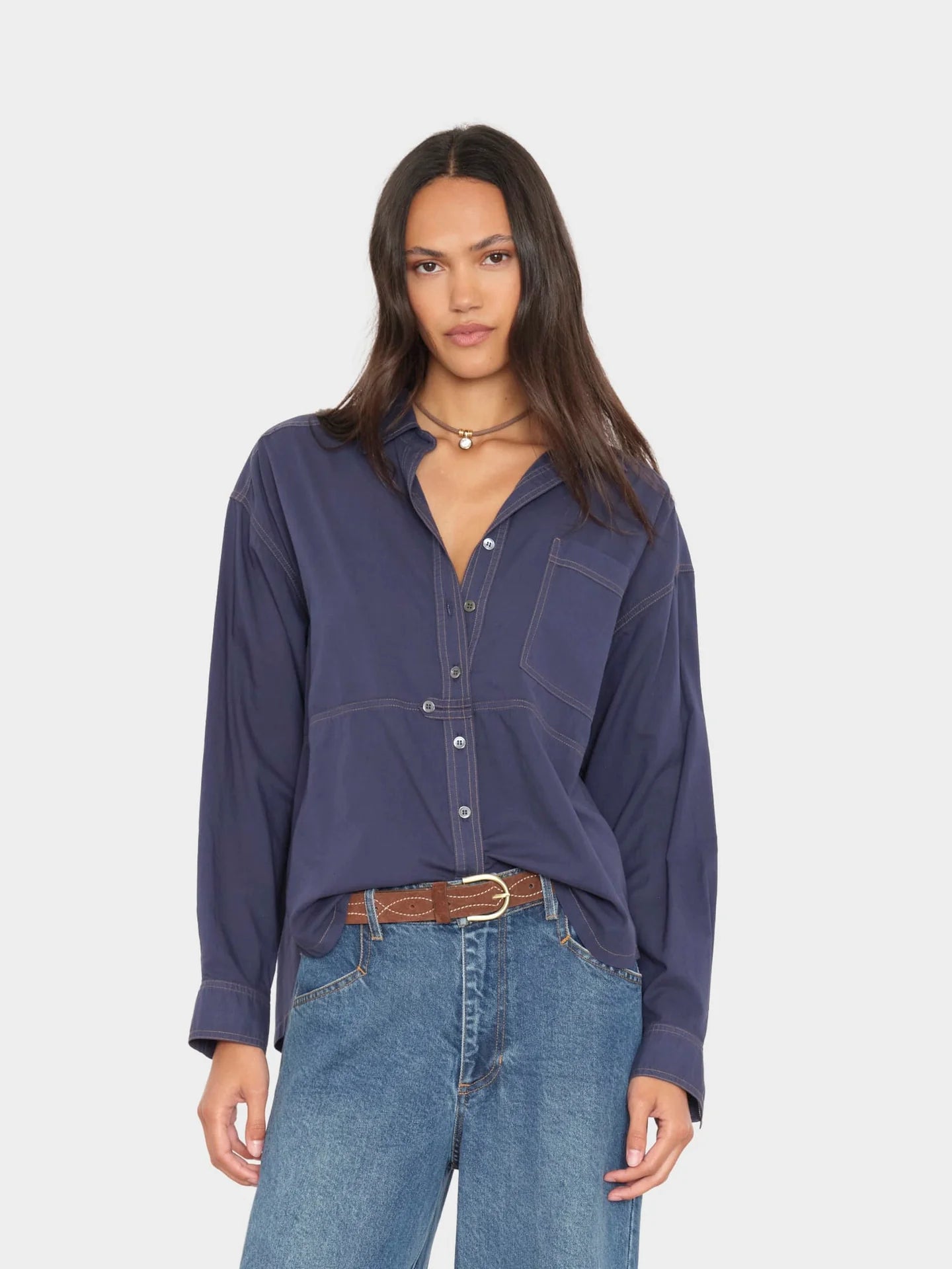 A woman modeling a dark blue long-sleeve button-up shirt with chest pockets, paired with blue jeans and a brown belt, against a light gray background.