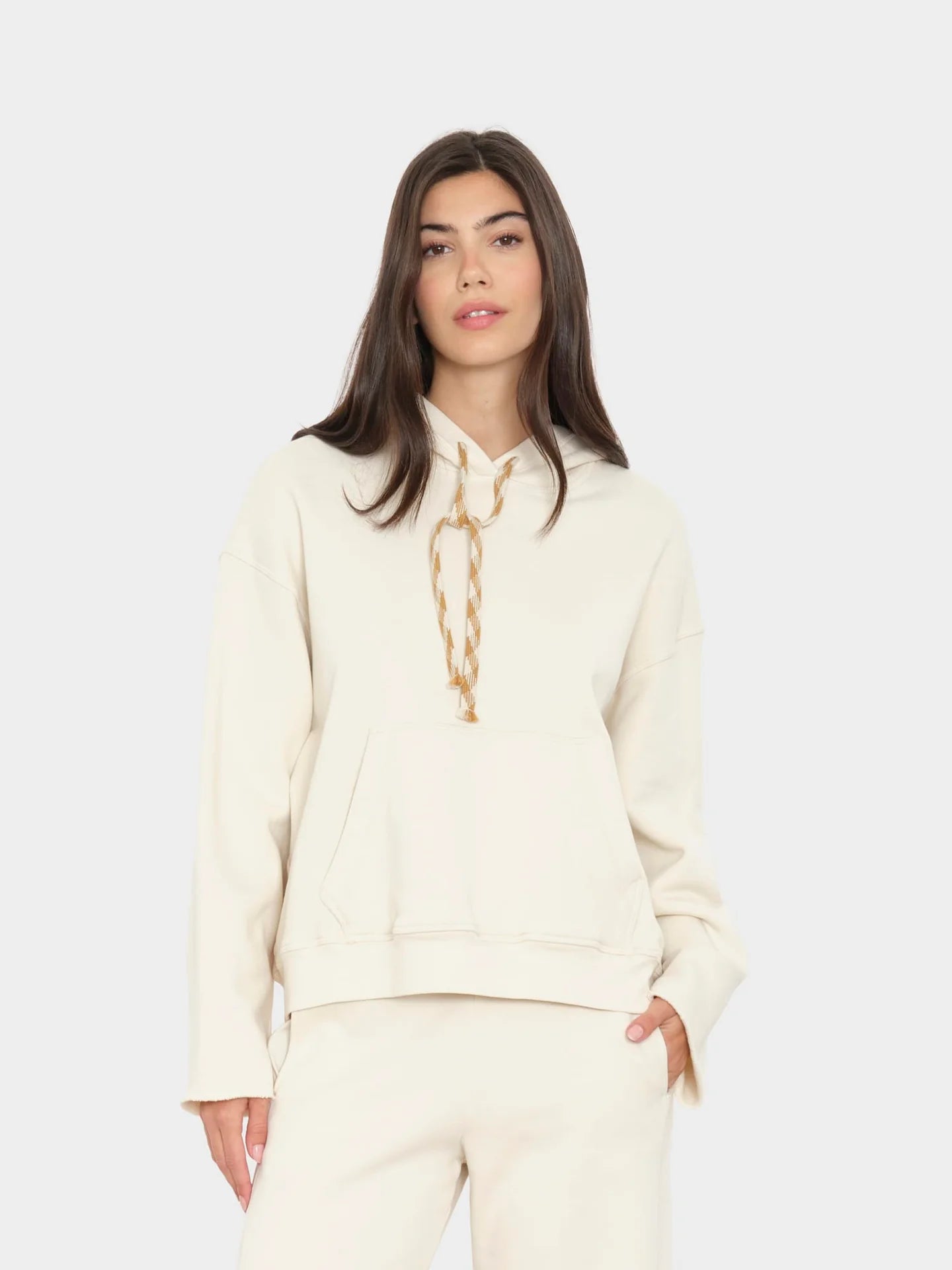 A woman modeling a cream-colored hoodie with a gold chain detail around the neck, paired with matching cream pants, against a light gray studio background.