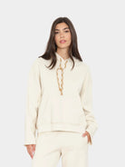 A woman modeling a cream-colored hoodie with a gold chain detail around the neck, paired with matching cream pants, against a light gray studio background.