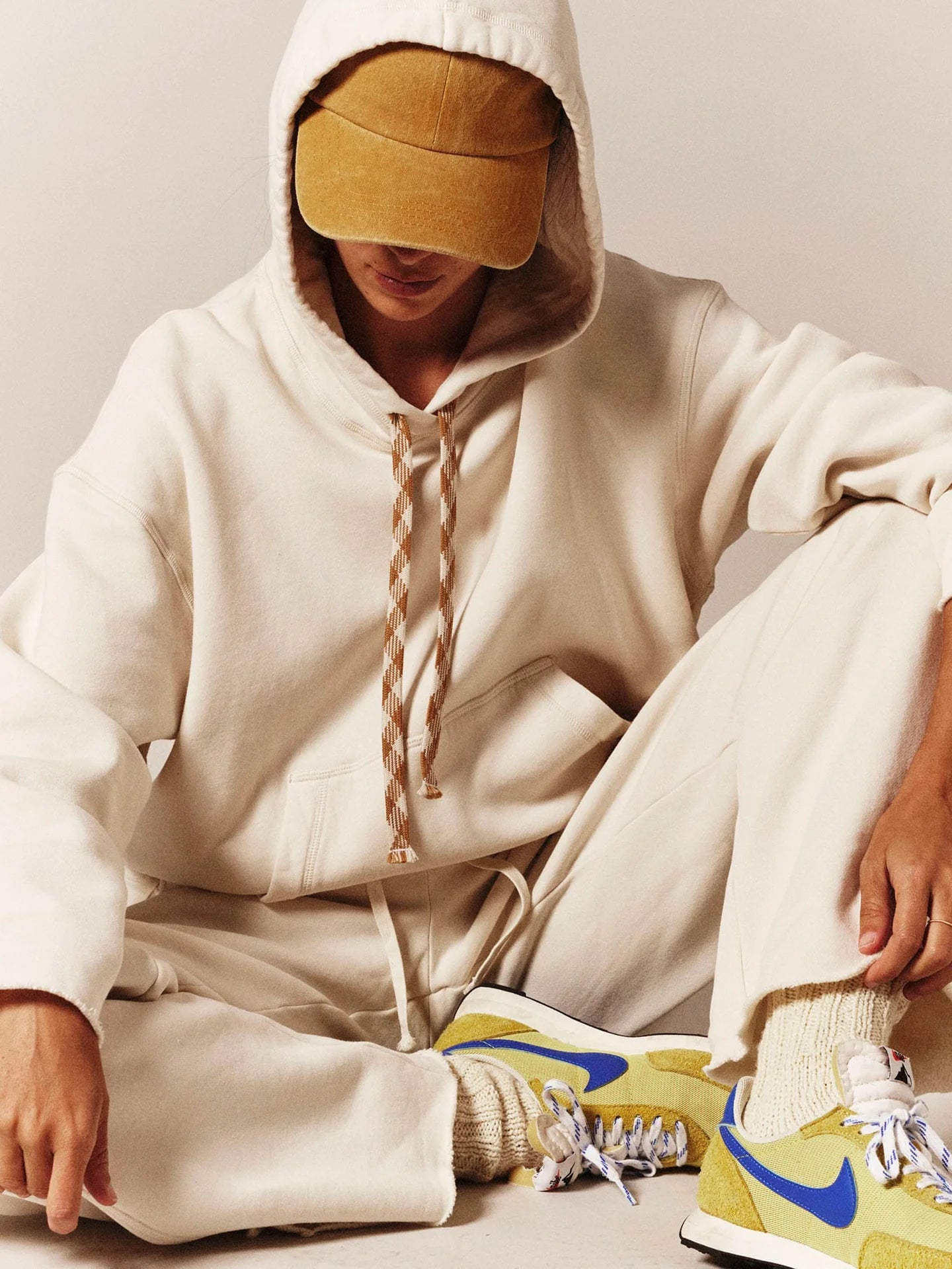 A person wearing a cream-colored hooded sweatshirt and matching sweatpants, seated on a light floor. They are wearing a mustard yellow cap and yellow sneakers with blue and white accents.